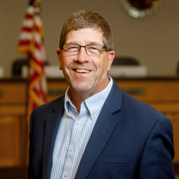 Man in blue suit posing with American flag and City seal in background